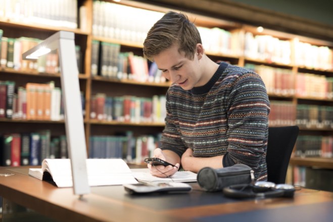 Gutt sitter ved en pult på biblioteket og ser på mobilen. foto